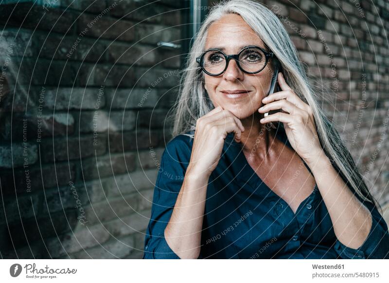 Smiling woman with long grey hair on cell phone on the phone call telephoning On The Telephone calling females women brick wall smiling smile mobile phone
