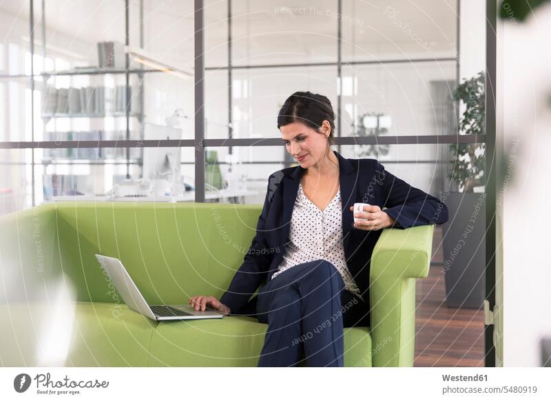 Businesswoman with laptop sitting on a couch and drinking coffee in the office caucasian caucasian ethnicity caucasian appearance european three-quarter length