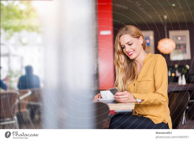 Smiling young woman sitting in a coffee shop looking at smartphone females women cafe Smartphone iPhone Smartphones Adults grown-ups grownups adult people