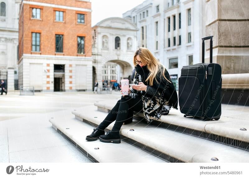 UK, London, young woman with luggage checking her cell phone on stairs paper cup papercups paper cups text messaging SMS Text Message City Break City Trip