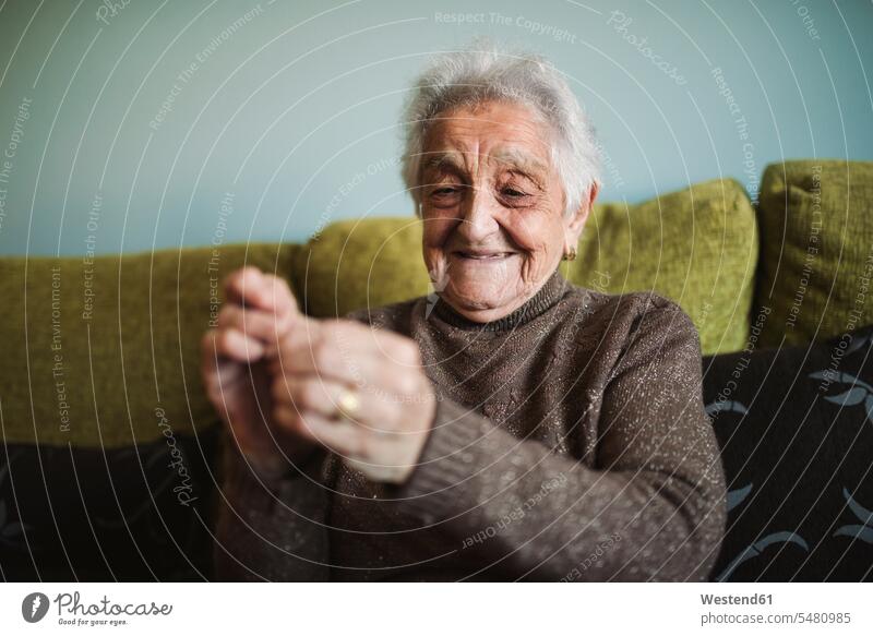 Portrait of smiling senior woman sitting on couch passing thread through buttonhole of sewing needle senior women elder women elder woman old portrait portraits