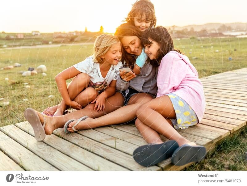 Four girls sitting together on boardwalk looking at cell phone females female friends Smartphone iPhone Smartphones child children kid kids people persons