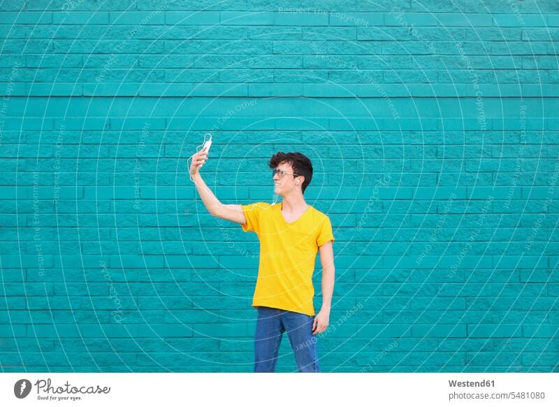 Young man taking a selfie with smartphone in front of blue brick wall men males Selfie Selfies Adults grown-ups grownups adult people persons human being humans