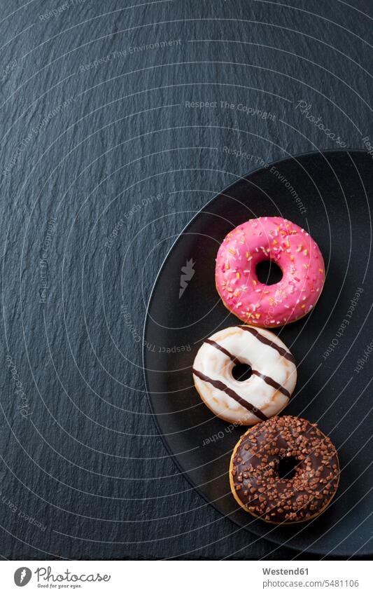 Three doughnuts with different icings on black plate and slate arrangement grouping sweet Sugary sweets round roundings donuts Doughnuts baked goods pastries