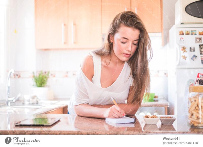 Young woman writing on notepad in kitchen pads Note Pad notepads Note Pads write females women Adults grown-ups grownups adult people persons human being humans