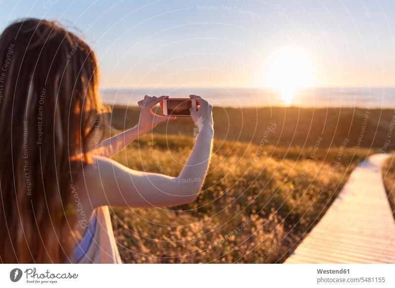 Young woman photographing beach at sunset with smart phone mobile phone mobiles mobile phones Cellphone cell phone cell phones Smartphone iPhone Smartphones