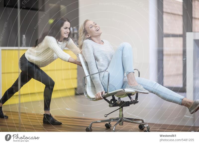 Young woman pushing her friend sitting on svivel chair through the office swivel chair office chair office chairs Office Offices Female Colleague colleagues