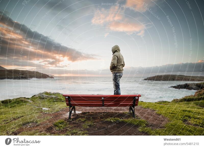 Spain, Ferrol, man wearing hooded jacket standing on a bench at the coast looking at distance coastline shoreline outdoors outdoor shots location shot
