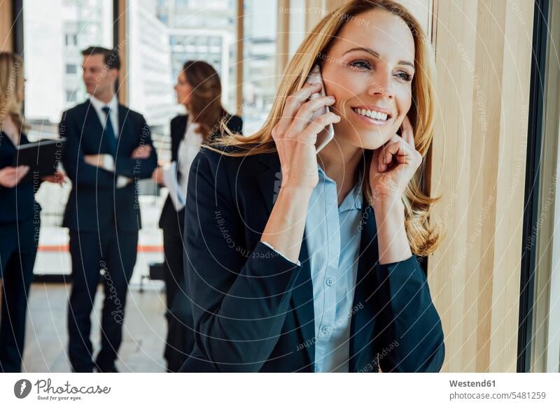 Smiling businesswoman in office on cell phone with businesspeople in background businesswomen business woman business women on the phone call telephoning