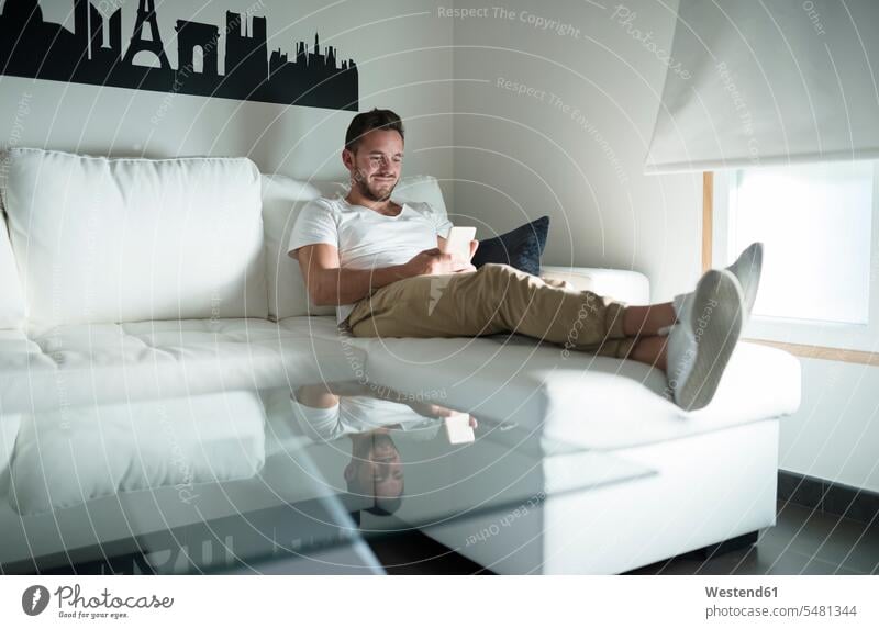 Smiling young man relaxing at home with his smartphone smiling smile couch settee sofa sofas couches settees sitting Seated mobile phone mobiles mobile phones