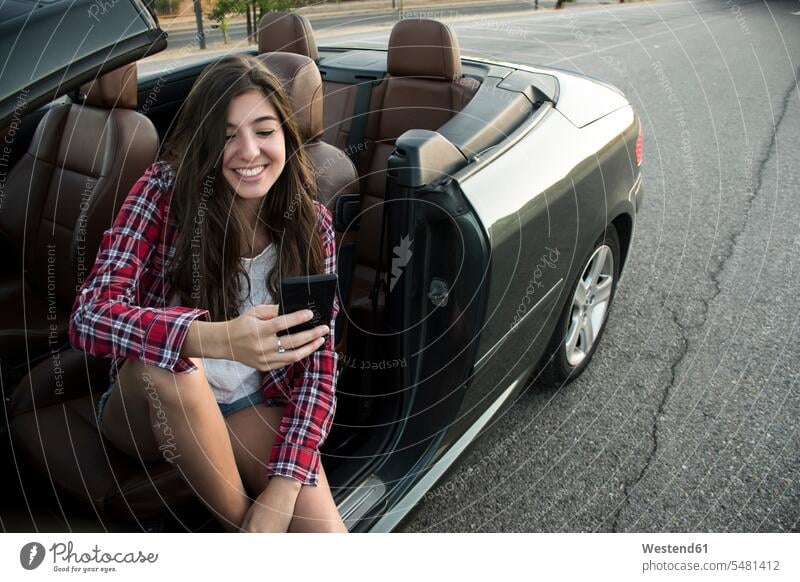 Smiling young woman sitting in convertible looking at smartphone females women smiling smile Seated car automobile Auto cars motorcars Automobiles motor vehicle
