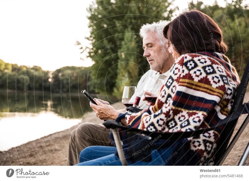 Senior couple with wine sitting at a lake with tablet lakes relaxed relaxation twosomes partnership couples digitizer Tablet Computer Tablet PC Tablet Computers