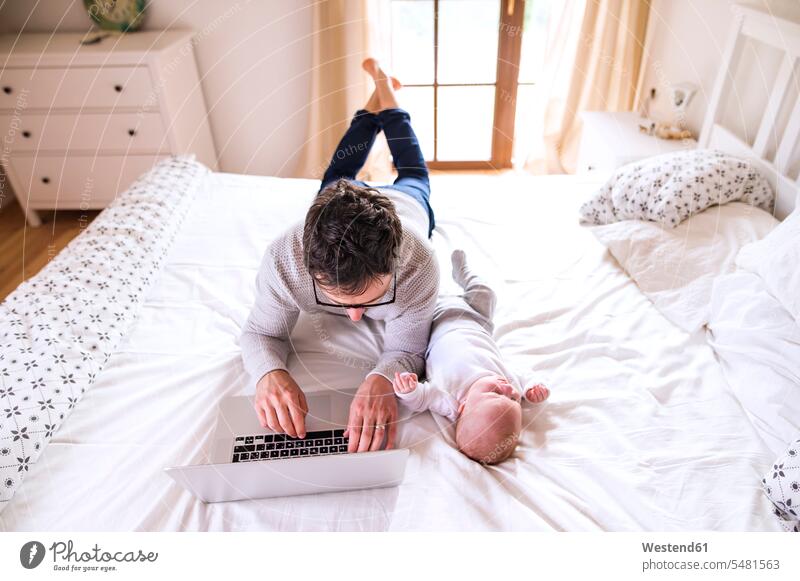 Father with baby lying on bed using laptop Laptop Computers laptops notebook father pa fathers daddy dads papa child children babies infants computer computers