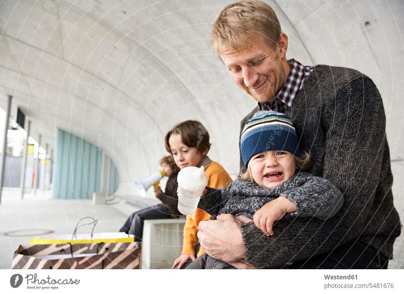 Crying boy sitting on father's lap, brother waiting in background megaphone megaphones bullhorns son sons manchild manchildren baby boys male Seated family
