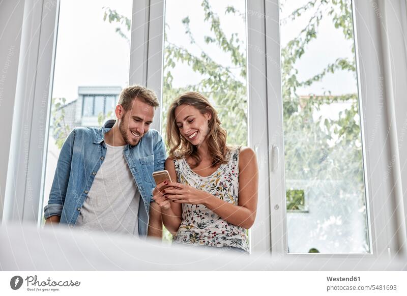 Smiling couple in front of window looking at smartphone twosomes partnership couples Smartphone iPhone Smartphones smiling smile people persons human being