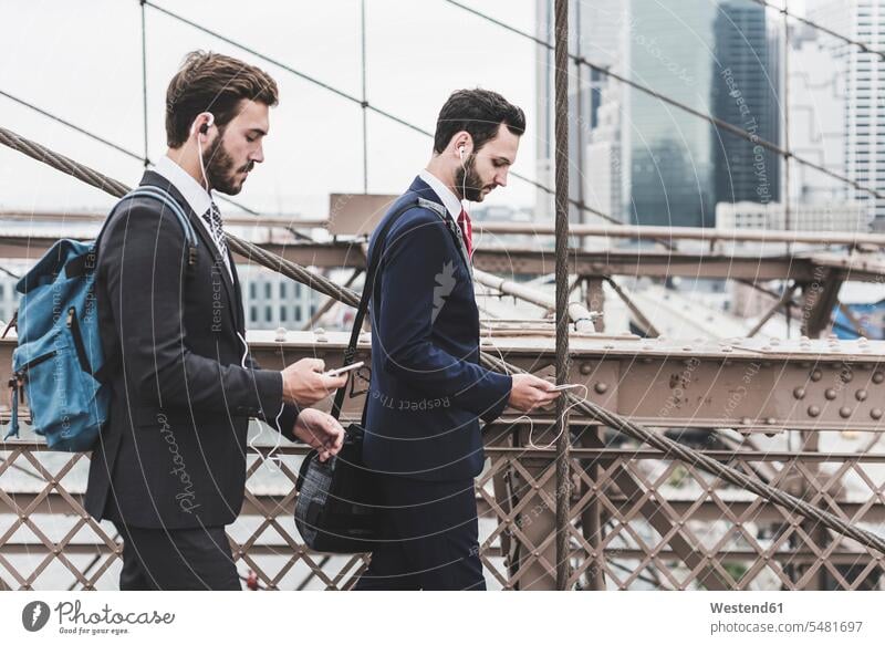 USA, New York City, two businessmen with cell phones and earbuds on Brooklyn Bridge Businessman Business man Businessmen Business men bridge bridges