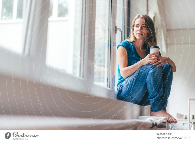 Smiling young woman holding takeaway coffee sitting at the window Coffee females women smiling smile relaxed relaxation Drink beverages Drinks Beverage
