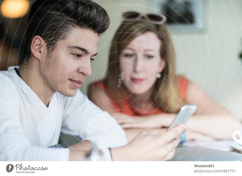 Teenage boy using cell phone with woman in background refugee refugees coffee shop cafe Teenager Teens teenagers godmother helper portrait portraits assistance