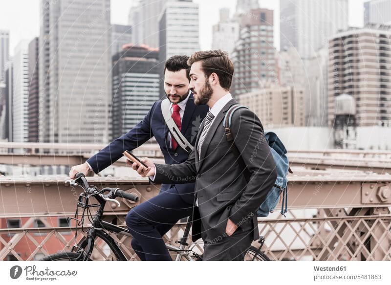 USA, New York City, two businessmen with bicycle and cell phone on Brooklyn Bridge talking speaking New York State Businessman Business man Businessmen
