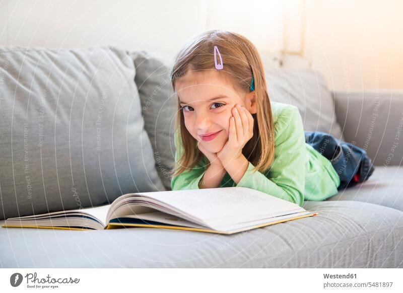Portrait of smiling little girl lying on couch with a book females girls books portrait portraits child children kid kids people persons human being humans
