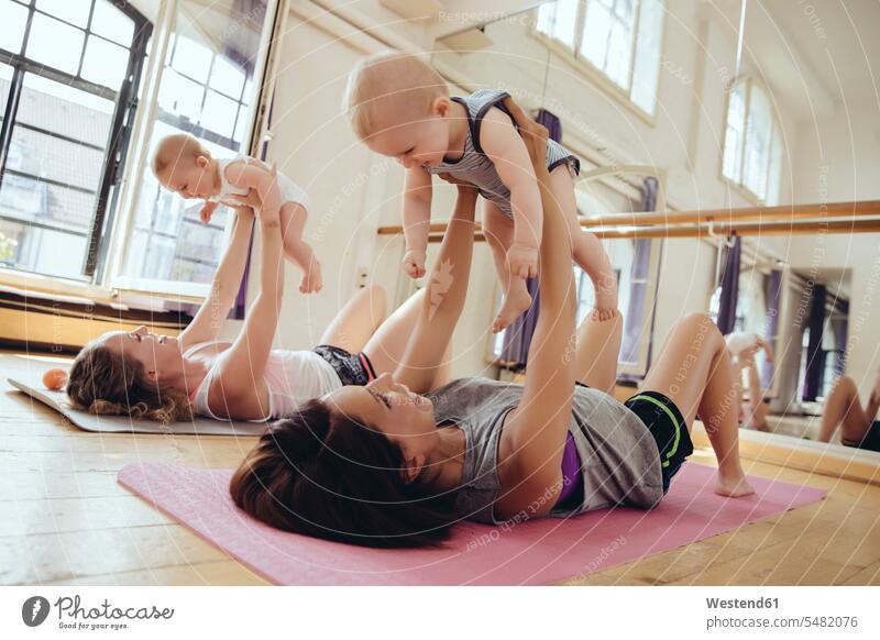 Two mothers working out on yoga mats while holding up their babies mommy mummy mama smiling smile lifting Fun having fun funny baby infants nurselings parents