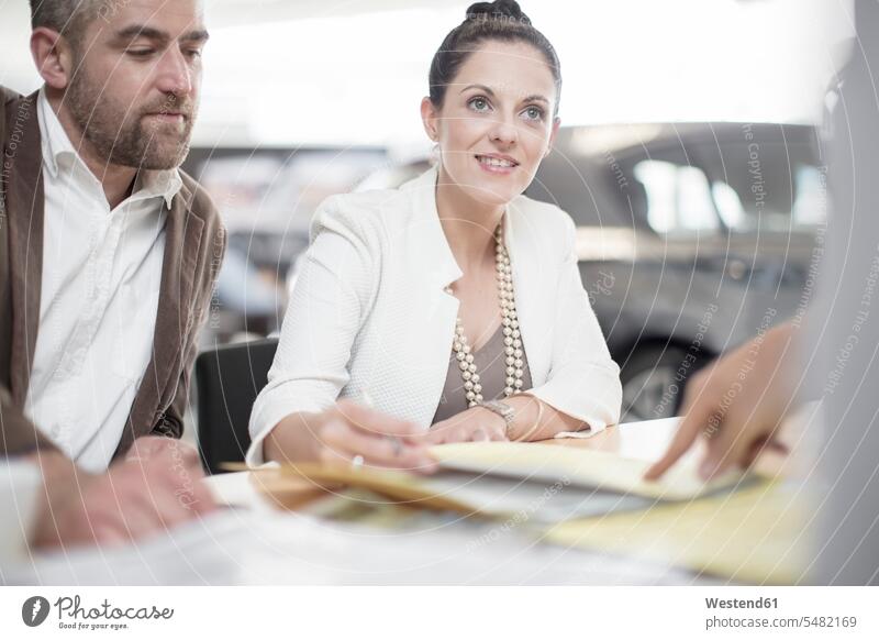 Car dealer and couple sitting at desk with agreement forms in showroom car dealer car dealers car dealership car dealerships twosomes partnership couples