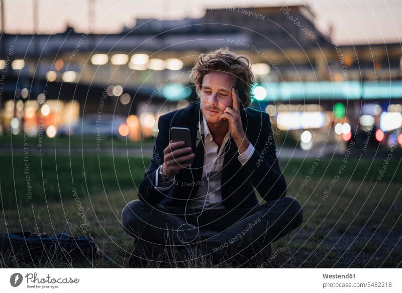 Businessman sitting on meadow at dusk with cell phone and earphones Business man Businessmen Business men mobile phone mobiles mobile phones Cellphone