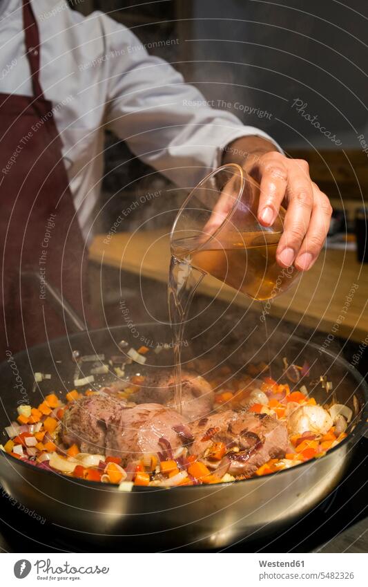 Man pouring a glass of brandy on beef cheeks in a pan with sauteed vegetables cooking chef cooks Chefs frying roasting Frying pan frying pans man men males