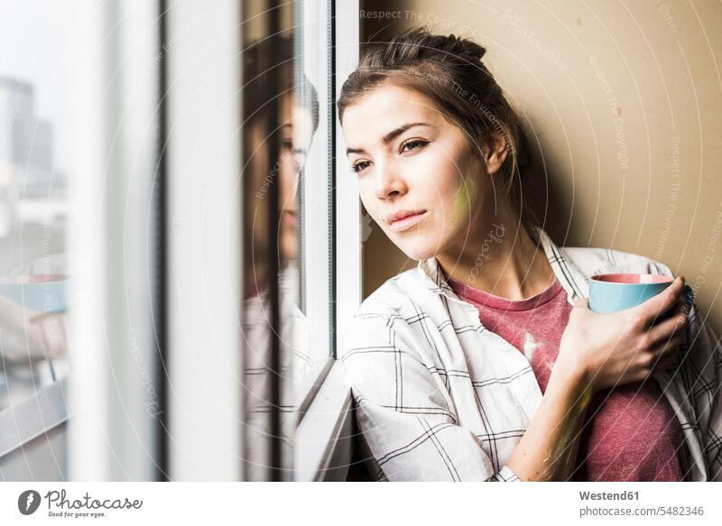 Young woman holding coffee cup, looking out of window young women young woman home ownership private owned home renovation renovate refurbish refurbishing