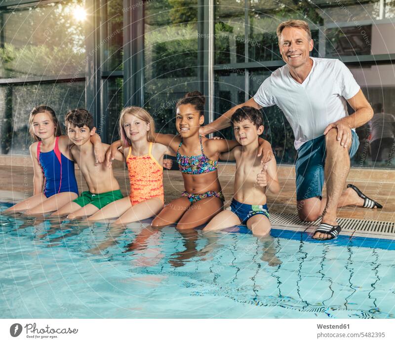 Portrait of smiling children with instructor sitting on poolside in indoor swimming pool coach coaches trainer portrait portraits pool edge Pool Side Seated