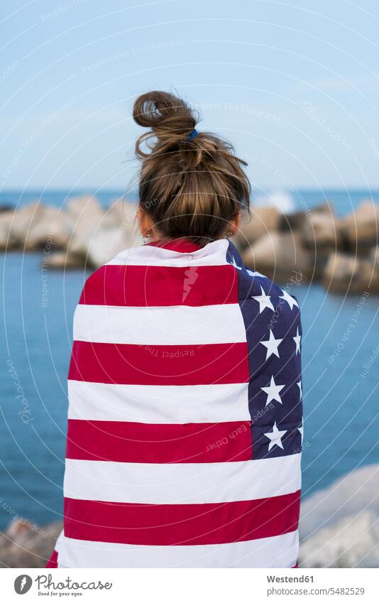 Back view of young woman standing in front of the sea wrapped in US American flag females women Adults grown-ups grownups adult people persons human being
