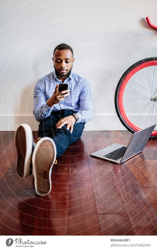 Man sitting on wooden floor with laptop and cell phone and bicycle next to him Businessman Business man Businessmen Business men Seated mobile phone mobiles