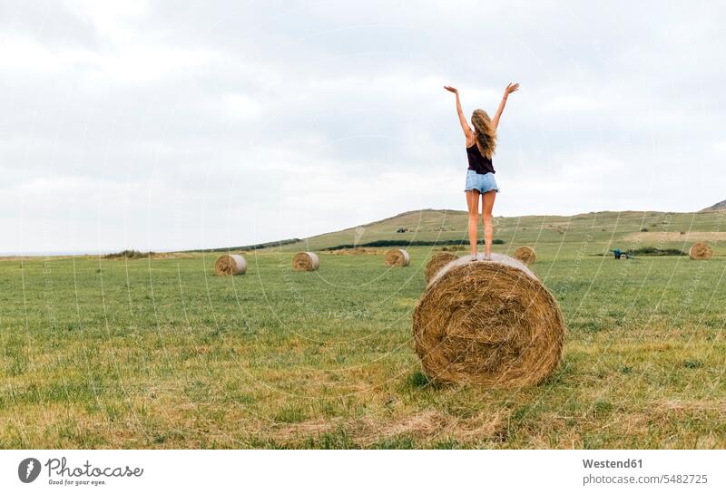 Back view of teenage girl standing on straw bale on a meadow raising arms Teenage Girls female teenagers Teenager Teens people persons human being humans