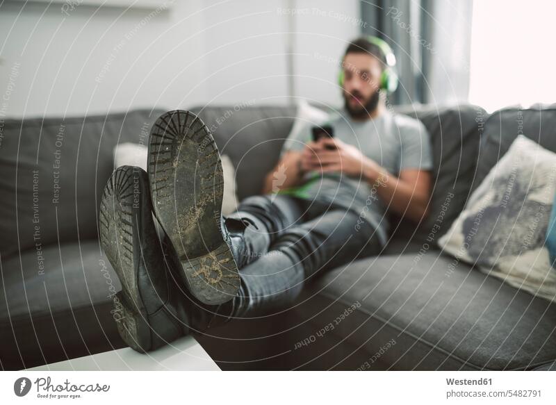 Young man sitting on sofa, listening music from his smart phone young men males Listening Music headphones headset mobile phone mobiles mobile phones Cellphone