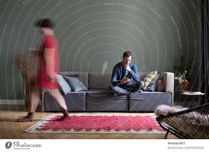 Man sitting on the couch in the living room while his girlfriend passing in the foreground Smartphone iPhone Smartphones man men males living rooms livingroom
