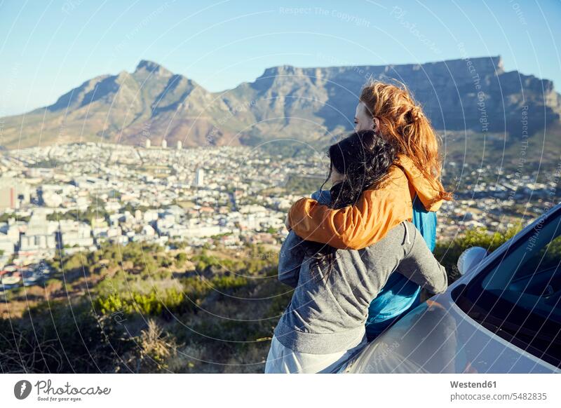 South Africa, Cape Town, Signal Hill, two young women leaning against car overlooking the city standing woman females embracing embrace Embracement hug hugging