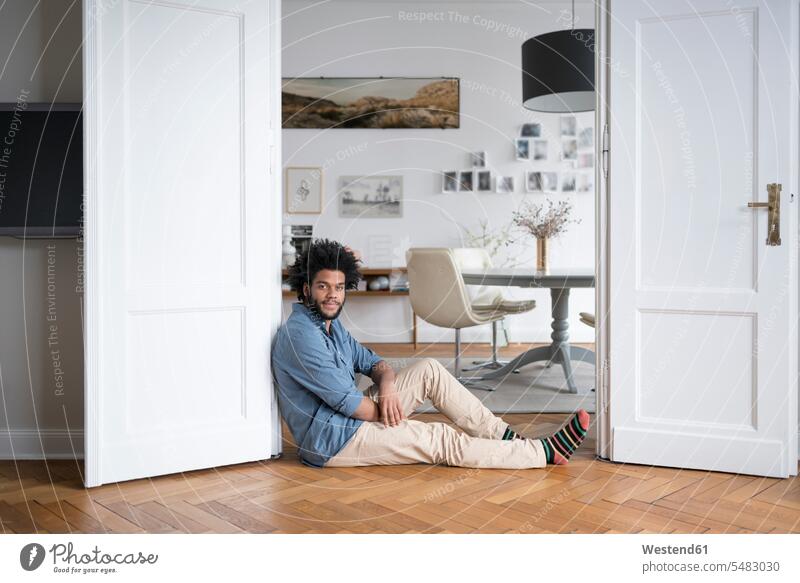 Man at home sitting on floor leaning on door frame in living room man men males smiling smile Adults grown-ups grownups adult people persons human being humans