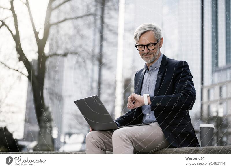 Grey-haired businessman working with laptop on wall in city looking at his smartwatch smiling smile wrist watch Wristwatch Wristwatches wrist watches