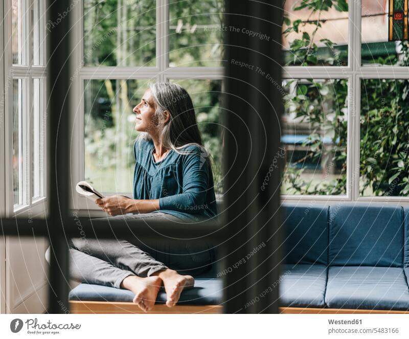 Relaxed woman sitting with book on lounge in winter garden looking through window females women winter gardens conservatory Sun Room Sunroom Adults grown-ups