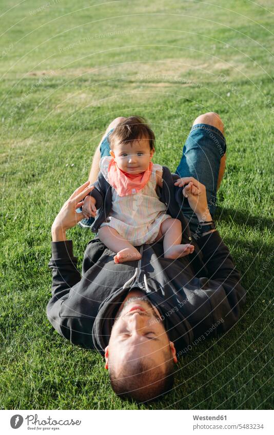 Cute baby girl sitting on her father's belly in a meadow relaxed relaxation pa fathers daddy dads papa smiling smile infants nurselings babies relaxing parents