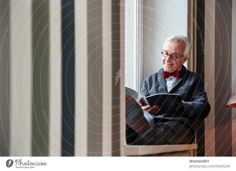 Senior man sitting in windowsill reading magazine senior men senior man elder man elder men senior citizen Seated window sill sills window sills Window Cill