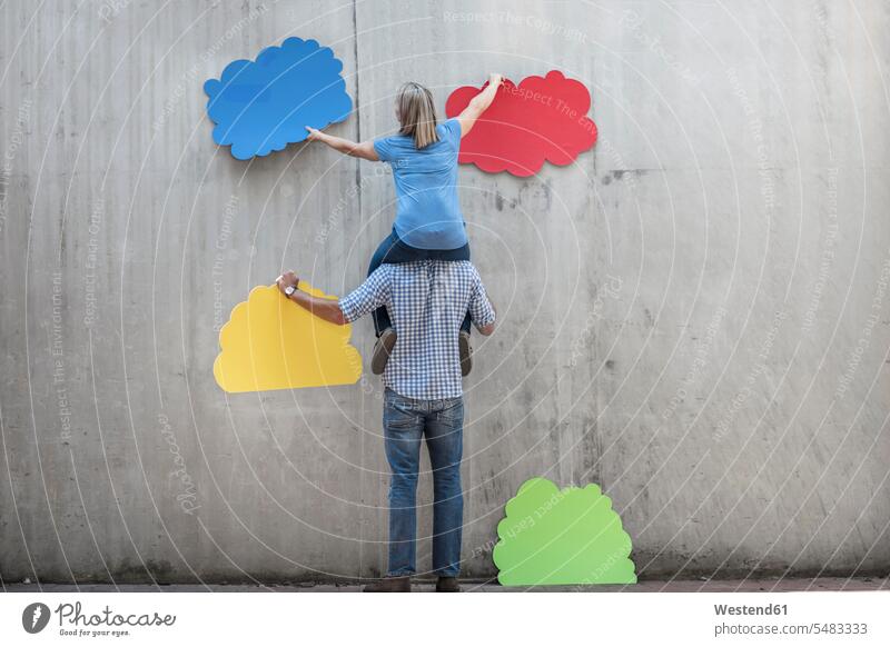 Woman sitting on a man's shoulders attaching colourful cloud shapes to concrete wall concrete walls multi-coloured multicoloured multi colored Multi Coloured