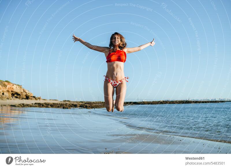 Smiling young woman jumping in the air on the beach beaches jump in the air females women Leaping jumps Adults grown-ups grownups adult people persons