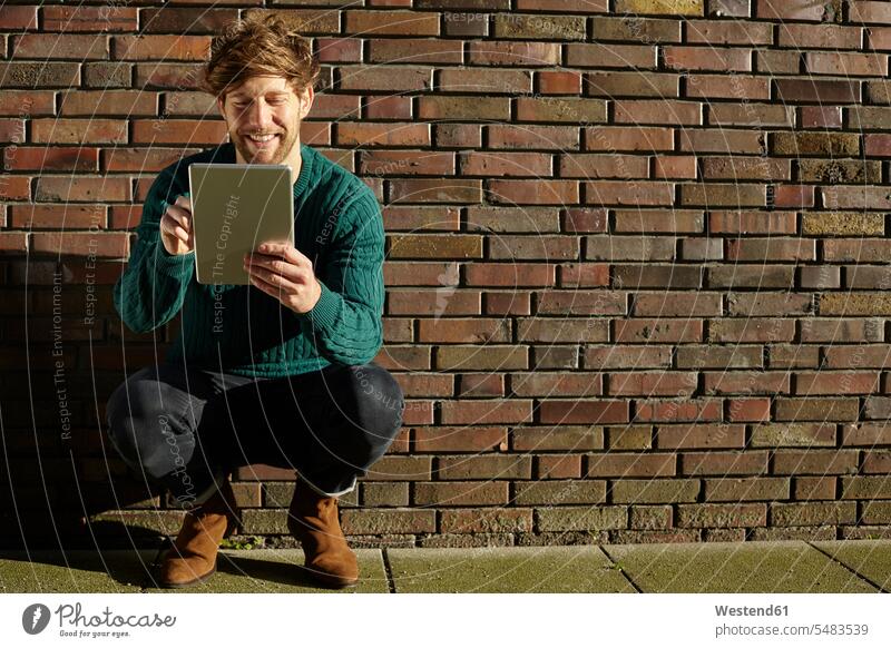 Young man crouching in front of a brick wall using digital tablet caucasian caucasian ethnicity caucasian appearance european Joy enjoyment pleasure Pleasant
