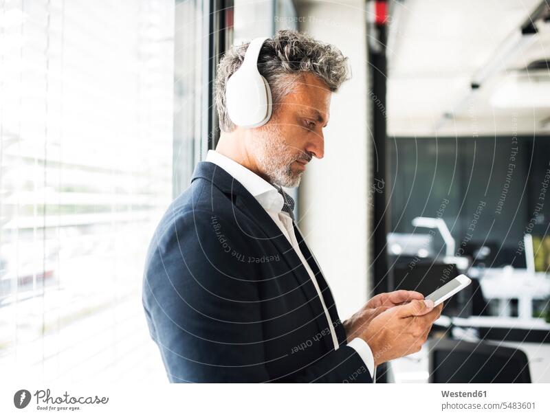 Mature businessman at the window in office with headphones and cell phone offices office room office rooms windows Businessman Business man Businessmen