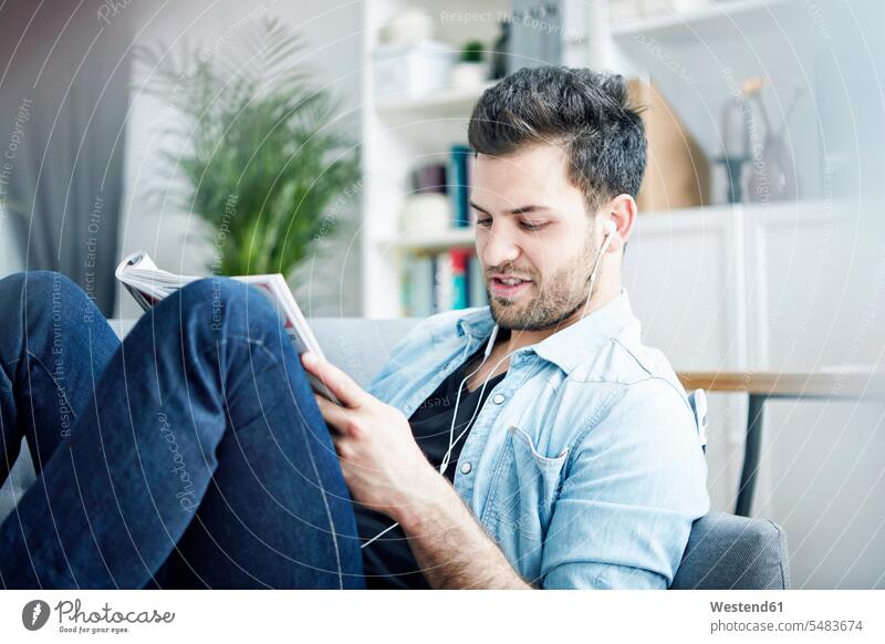 Young man at home with earbuds reading magazine living room living rooms livingroom headphones headset shirt shirts relaxation relaxed relaxing stubble Unshaven