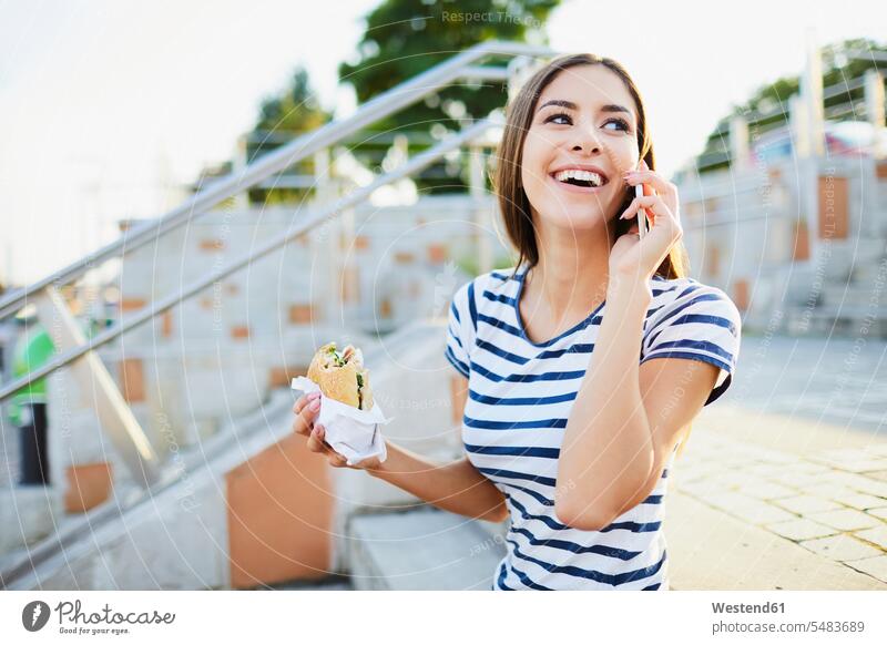 Young woman talking on phone and eating bagel in the city on the phone call telephoning On The Telephone calling mobile phone mobiles mobile phones Cellphone