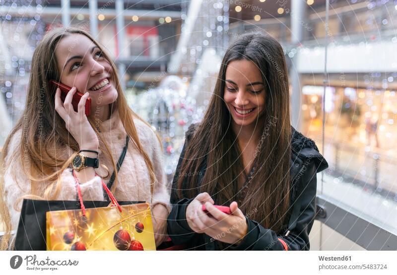 Two female friends with shopping bags on an escalator of a shopping center using their smartphones paper bag paper bags toothy smile big smile open smile