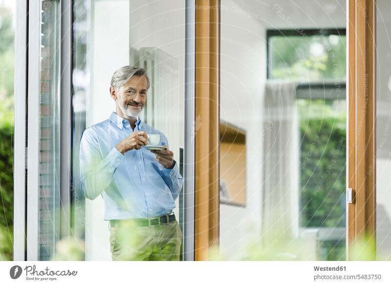 Smiling man with cup of coffee standing in his house looking through window men males Adults grown-ups grownups adult people persons human being humans