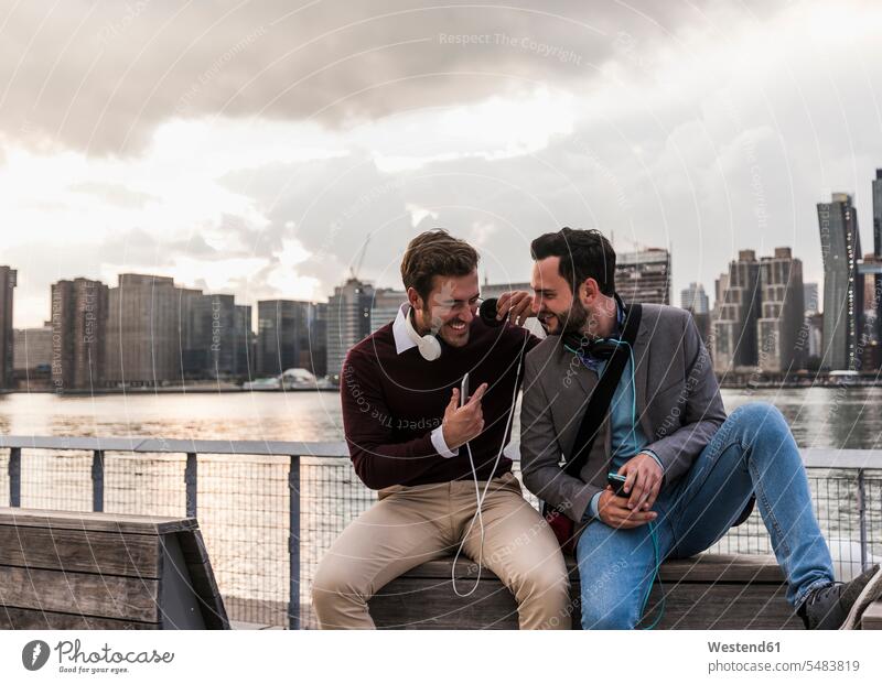 USA, New York City, two happy young men with headphones and cell phone sitting at East River laughing Laughter friends New York State man males positive Emotion